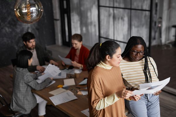 Duas mulheres no primeiro plano e mais um time de 3 pessoas ao fundo representando times de RH com papel estratégico, trabalhando na reestruturação de times e mudanças em empresas.